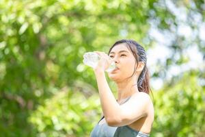 deportista Bebiendo agua después trabajando fuera hacer ejercicio, atleta joven mujer con un el plastico agua botella a verde parque foto