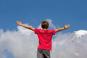 A tourist boy stands on the top of a hill with his arms spread wide to the sides against the blue sky. Travel concept, freedom. Moving forward towards the unknown. Freedom of choice. photo