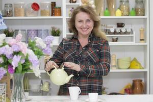 hermosa contento ordinario mayor mujer en el cocina con un tetera y un taza foto