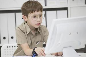 un niño con lentes a el computadora. colegial mira a el monitor pantalla foto