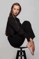 A slender beautiful teenage girl in a black jacket sat in a beautiful pose on a studio chair. Blonde fashion model girl posing. The dream is to be a model. photo