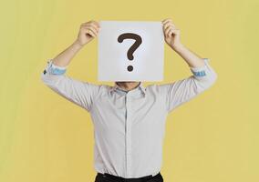 Young businessman covering face holding paper with question mark symbol. Standing over isolated yellow background photo