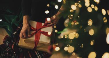 Hands in cozy sweater opening christmas gift with red bow on background of christmas tree with lights. Stylish female holding present with red ribbon in festive room close up. Merry Christmas photo