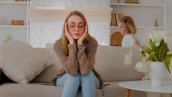 Tired upset Caucasian mother sitting on sofa at home sad frustrated stressed exhausted fatigued mom suffer headache with active noise difficult kid child baby girl daughter running around playing photo