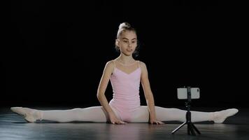 Girl teenager child ballerina acrobat gymnast sits on floor dance class online lesson with Internet coach trainer teacher remotely with smartphone on tripod sitting on twine recording video vlog blog photo
