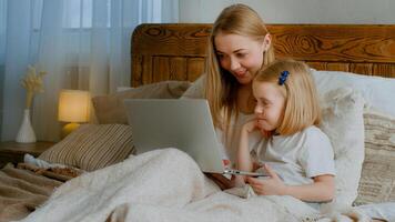 Caucasian woman mother mom with little child daughter baby girl preschool kid lying in bed at home bedroom with laptop using computer watching movie cartoons browsing together online shopping family photo