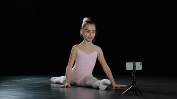 talentoso bailarina niña gimnasta acróbata se sienta en enroscarse haciendo espalda curva dispara vídeo Blog vlog en línea lección en móvil teléfono cámara teléfono inteligente en trípode acrobático elemento aprender con Internet web entrenador foto