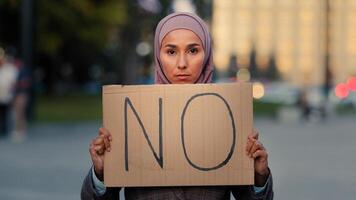 Stop racism No concept Arab immigrant Muslim woman in hijab protests against discrimination vax vaccination standing in city. Islamic girl holding cardboard slogan banner with text no disagree refusal photo