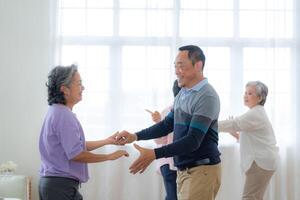 Asian Older male and females people dance with their partners on a dancing floor in living space. Happy older couple performing get exercise. Joyful carefree retired senior friends enjoying relaxation photo