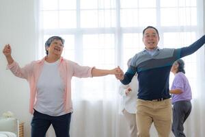 Asian Older male and females people dance with their partners on a dancing floor in living space. Happy older couple performing get exercise. Joyful carefree retired senior friends enjoying relaxation photo