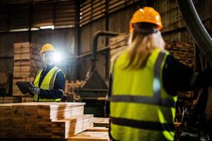 Team worker carpenter wearing safety uniform and hard hat working and checking the quality of wooden products at workshop manufacturing. man and woman workers wood in dark warehouse industry. photo