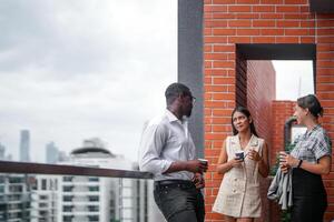 equipo de negocio personas inteligente hombre y mujer estar a al aire libre terraza edificio y hablar juntos con desayuno comida y café en el mano en bueno sensación con ciudad espacio edificio. negocio Mañana. foto