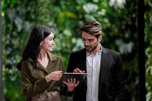 Businessman and casual businesswoman talking outdoors. Two young multicultural coworkers watching online video in tablet held by happy businesswoman. photo