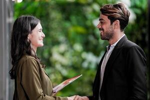 Businessman and casual businesswoman shaking hands outdoors. Two young multicultural coworkers deal for success. background of  green wall nature trees. photo