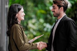 empresario y casual mujer de negocios sacudida manos al aire libre. dos joven multicultural compañeros de trabajo acuerdo para éxito. antecedentes de verde pared naturaleza arboles foto