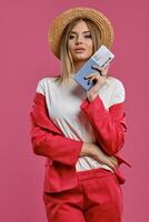 Blonde model in straw hat, white blouse and red pantsuit. She is holding passport and ticket while posing against pink studio background. Close-up photo