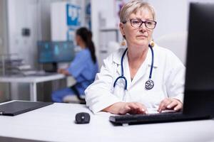 Expert woman doctor working at computer, entering data from patient file. Female physician in white coat and stethoscope sitting in hospital office typing on pc keyboard with nurse in background photo
