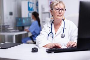 Expert woman doctor working at computer, entering data from patient file. Female physician in white coat and stethoscope sitting in hospital office typing on pc keyboard with nurse in background photo