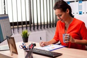 Professional businesswoman entrepreneur reading finance analysis looking at corporate charts wearing red. Successful financial auditor in data marteking looking at graphs on computer screen and writing report. photo