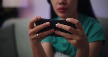 metraggio vicino su di giovane asiatico donna indossare cuffie gode giocando in linea esport Giochi su smartphone seduta su divano nel il vivente camera a casa, giocatore stile di vita concetto. video