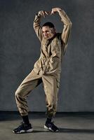 Active guy with tattooed body and face, earrings, beard. Dressed in khaki overalls and black sneakers. Dancing on gray background. Dancehall, hip-hop photo