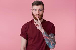 Poptrait of young bearded man with tattooed hand isolated over pink background, with wide open eyes, keeps fore finger on lips, keep quiet, do not make noise, demonstrates silence gesture. photo