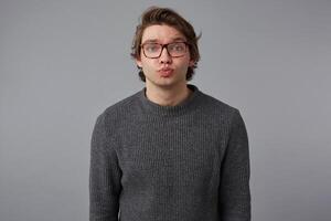 Portrait of young man with glasses wears in gray sweater, stands over gray background and sends kiss to the camera. photo