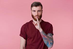 Close up of bearded young man with tattooed hand keeps fore finger on lips, calls to keep a secret, do not tell anyone, do not make noise, demonstrates silence gesture, isolated over pink background. photo