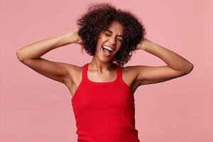 bonito joven africano americano niña disfruta bailando con su ojos cerrado, toques su Rizado afro cabello, sostiene su manos a su cabeza, abrió boca como cantando, tiene divertido, vistiendo rojo camiseta, aislado en rosado foto