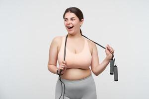 Studio photo of glad young dark haired chubby woman with jumping-rope on her neck looking cheerfully aside with wide smile, going to make sports while posing over white background