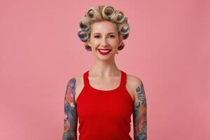 Cheerful young blonde tattooed female with evening makeup making hairdo while posing over pink background with hands down, looking at camera with charming smile photo