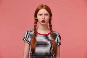 Concerned and worried young 20s woman with two red haired braids red lipstick open mouth in despair and shock not knowing what do standing troubled facing huge problem upset and puzzled over pink wall photo