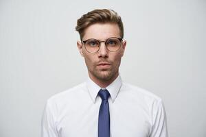 Photo as on passport. Young man with stylish haircut, glasses,blue eyes look straight serious and self-collected, dressed in white shirt and blue tie, unshaven, over white background