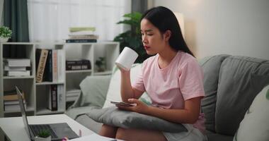 beeldmateriaal van jong Aziatisch vrouw drinken koffie terwijl gebruik makend van een rekenmachine Aan smartphone en maakt aantekeningen in een notitieboekje Aan de bank, thuis financiën video