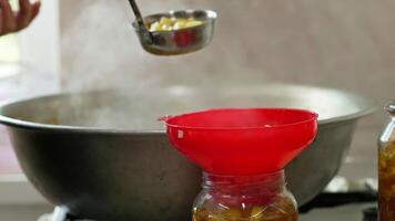 woman hands filling glass jars with apple jam via red funnel and soup ladle, slow motion closeup video