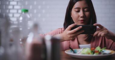 Footage of Young Asian woman sitting at a table enjoying a salad and use smartphone taking a photo in kitchen at home,Healthy food and wellness concept video