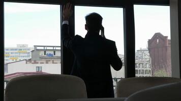 Troubled businessman in silhouette talking on the phone near the windows in his office. Dolly slider 4K footage video