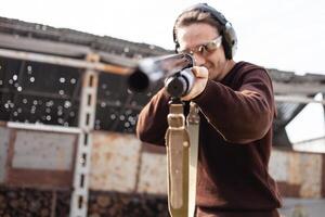 A man in protective glasses and headphones. A pump-action shotgun. Tyre's outdoors. A wall and a roof with bullet holes. Sport photo