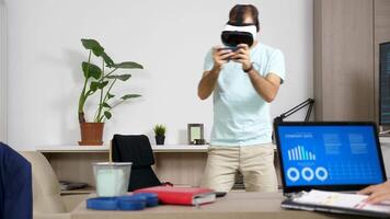 Man playing with VR headset while his girlfriend works on the computer in the same room. She looks at animated company data and chart. Rack focus dolly slider 4K footage video