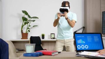 Man playing with VR headset while his girlfriend works on the computer in the same room. She looks at animated company data and chart. Rack focus dolly slider 4K footage video