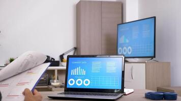 Back view of man looking at computer screen checking company data on animated charts. There is a big screen TV in background playing the same info. Dolly slider 4K footage with parallax effect video