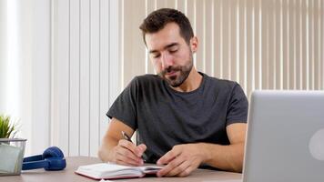 Mens Bij huis in gewoontjes kleren schrijven in een notebook. Daar is een laptop Aan de bureau hij is werken Aan. dolly schuif 4k beeldmateriaal video