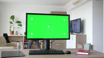 Personal PC with big green screen chroma mock up on the table in the living room. A man walks in the background while the TV is on and sits on the couch looking at his smartphone video