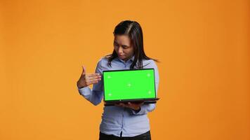 Confident adult showing laptop with greenscreen in studio, presenting isolated mockup template on wireless computer. Asian person holding pc with chromakey display on camera. video