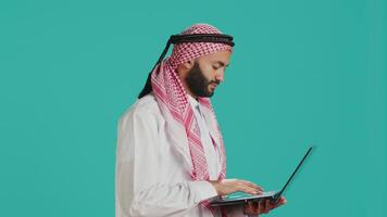 Person using laptop touchpad in studio, dressed in traditional muslim thobe and kufiyah. Middle eastern guy holding wireless computer and scrolling webpages or communicating on internet. video
