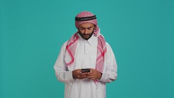 musulman homme navigation téléphone intelligent app et sentiment de bonne humeur après découverte en dehors à propos génial nouvelles, dactylographie des textes sur social médias site Internet. milieu est gars défilement en ligne pages, traditionnel vêtements. video