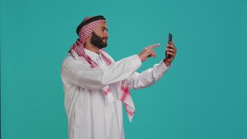 Arab man takes pictures with smartphone, laughing in studio using smartphone in front of studio camera. Middle eastern person with traditional headscarf taking photos in cultural attire. video