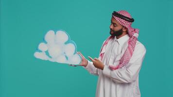 Middle eastern adult holds cloud icon on camera, showcasing blank billboard icon in natural layout. Male model in traditional muslim apparel and headdress doing new commercial advertisement. video