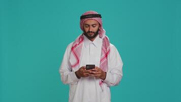 islamico tipo nel accappatoio sms su Telefono mentre in posa con fiducia al di sopra di blu sfondo. mezzo orientale persona vestito nel thobe e foulard, invio in linea messaggi su smartphone app. video