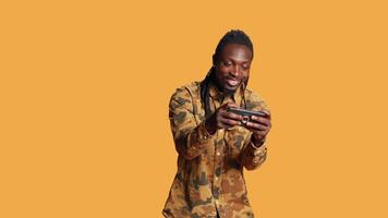 Joyful person playing online videogames on mobile phone, having fun with gaming tournament on device in studio. African american man enjoying shooting game with friends, orange background. video